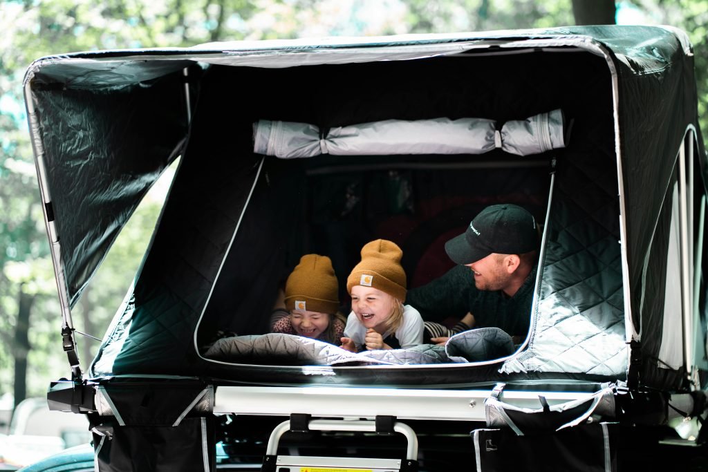 A father and his children enjoy laughter and togetherness in a rooftop tent during a summer camping trip in the woods.